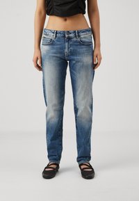 Egy nő elhalványult kék egyenes lábú farmert és fekete balettcipőt visel, egy sima világos háttér előtt áll.