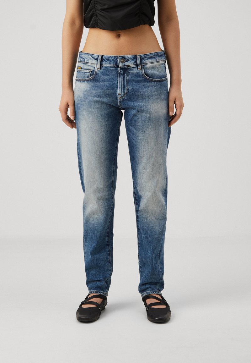 Egy nő elhalványult kék egyenes lábú farmert és fekete balettcipőt visel, egy sima világos háttér előtt áll.