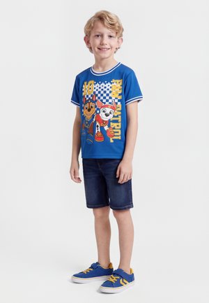 Smiling blonde boy standing wearing blue Paw Patrol t-shirt, dark denim shorts, and blue sneakers with yellow accents on a white background.
