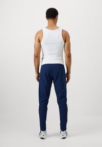 Homem de pé de costas, vestido com uma camisola branca, calças azul-marinho e ténis brancos, contra um fundo claro e liso.