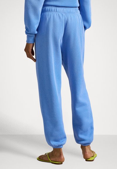Lyseblå joggebukser laget av mykt stoff, med elastisk midjebånd og mansjetter, med en avslappet passform, brukt sammen med grønne flip-flops.