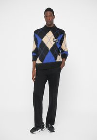 Man staat en draagt een blauw, zwart en beige argyle sweater met het logo "Versace Couture", zwarte broek en zwarte sneakers tegen een witte achtergrond.
