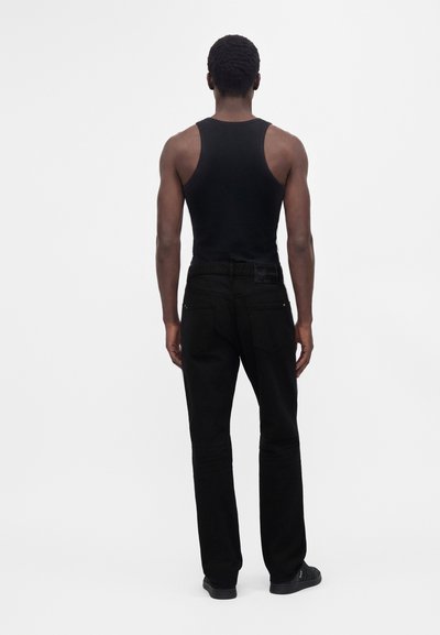 Homme portant un débardeur noir sans manches, un pantalon noir et des chaussures noires, debout de dos devant un fond clair uni.