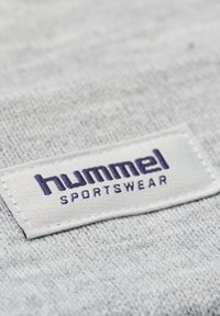 Grått tyg som visar en strukturstickning med en vit rektangulär etikett. Etiketten visar "hummel" i lila versaler, med "SPORTSWEAR" nedanför.