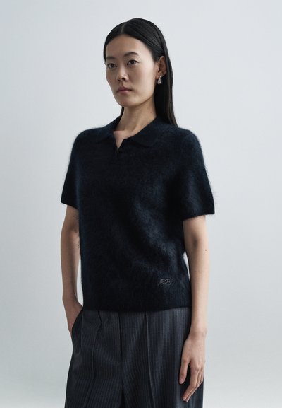 Pull polo à manches courtes en maille foncée, confectionné en matière douce et duveteuse. Présente un col et un détail de logo subtil à l'ourlet. Porté avec un pantalon rayé.