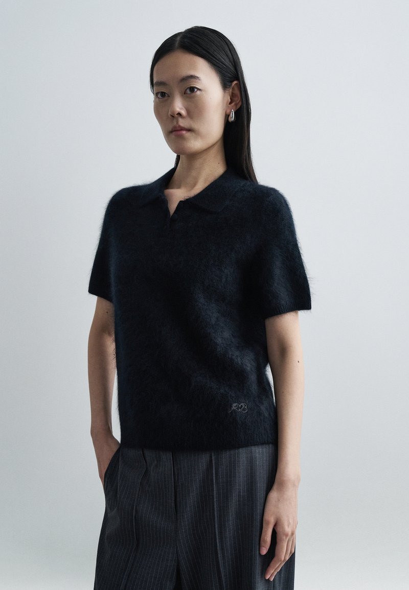 Mørk strikket polo-sweater med korte ærmer lavet af blødt, fluffy materiale. Har en krave og diskret logodetalje ved kanten. Stribede bukser båret.