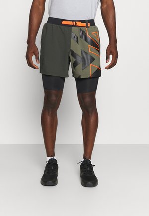 Un homme porte un short de sport superposé vert olive et noir avec des motifs géométriques orange et une ceinture orange vif, assorti à des chaussures de course noires.