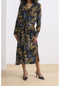 Robe noire à motif floral avec des manches longues, un nœud à l’avant et des fentes sur les côtés. Elle présente des fleurs jaunes et bleues, un tissu lisse et une coupe décontractée.