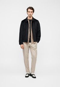Veste en daim noir portée sur une chemise beige à boutons et un t-shirt noir, associée à un pantalon beige clair et des mocassins noirs avec des accents blancs.
