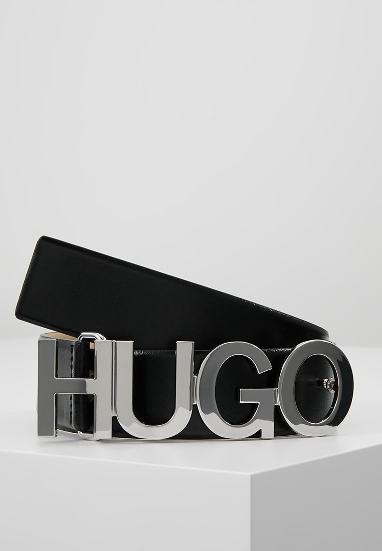 Ceinture en cuir noir avec une grande boucle argentée affichant "HUGO", posée sur une surface blanche.
