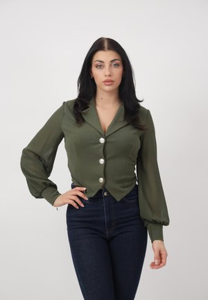 Kvinde med langt mørkt hår iført en olivengrøn skjorte med knapper og puffede ærmer samt mørkeblå bukser med høj talje, stående foran en ensfarvet baggrund.