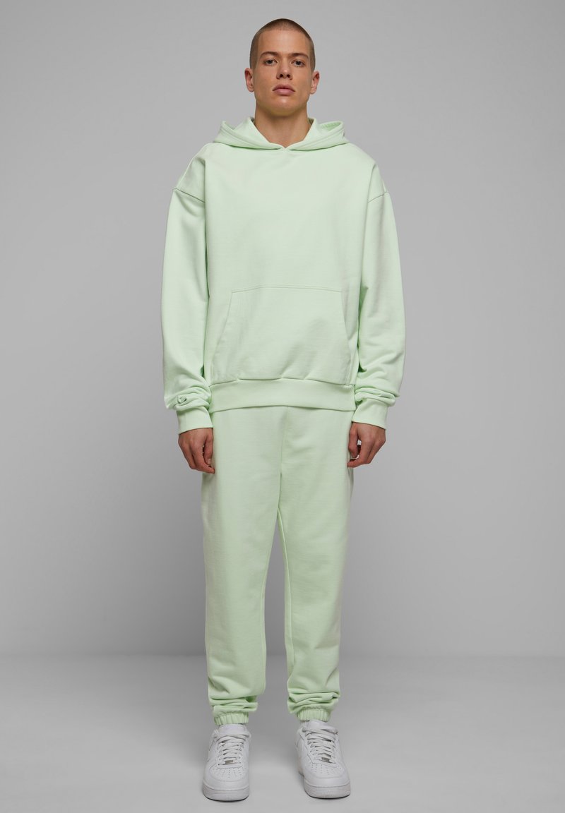 Sudadera de gran tamaño en verde claro, con bolsillo canguro, combinada con pantalones de chándal ajustados a juego. Presenta puños acanalados y zapatillas blancas.