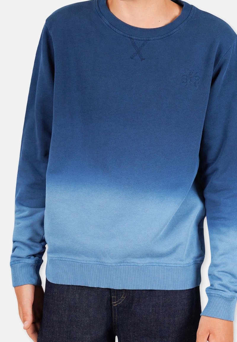 Hombre vestido con un sudadera de manga larga tipo crew neck con un degradado azul que va de oscuro en la parte superior a claro en la parte inferior, combinada con jeans oscuros.