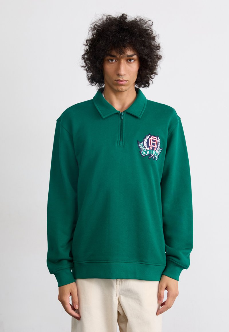 Sweatshirt verde com gola com zíper, mangas longas e um logótipo bordado com detalhes de louros no peito. Textura suave.