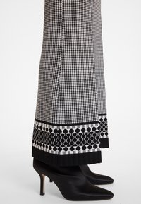 Schwarz-weiße gemusterte Hose mit Hahnentrittmuster; hat einen kontrastierenden hexagonalen Druck am Saum; kombiniert mit schwarzen Stilettos.