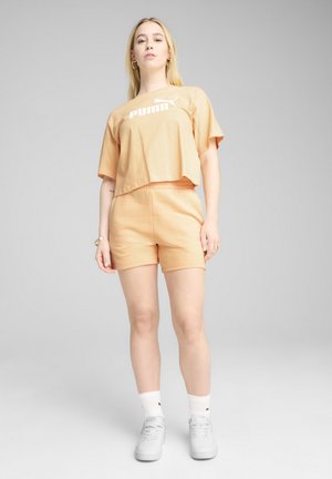 Šortky v broskyňovej farbe a zodpovedajúce skrátené tričko s bielym logom. Materiál sa zdá byť mäkký. Model stojí na neutrálnom pozadí.