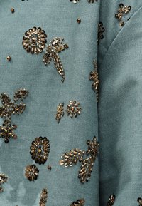 Mėlyna audinys su žąsies kaulo raštu, papuoštas išblaškytomis bronzos spalvos blizgančiomis dygsnelėmis ir karoliukais, sudarančiais abstrakčias gėlių formas.