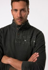 Suéter de color gris oscuro con un cuello alto, dos botones marrones y detalles texturizados. Presenta letras impresas en el pecho. Tejido suave.
