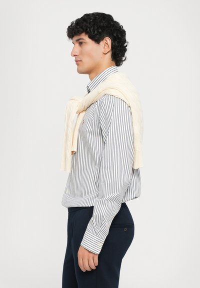 Chemise à rayures blanches et marines à manches longues, associée à un pantalon marine. Un pull en tricot cable de couleur jaune pâle est drapé sur les épaules.
