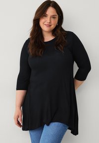 Sort langærmet tunika med rund halsudskæring og buet kant. Lavet af blødt stof, hænger den løst over kroppen og er parret med blå jeans.