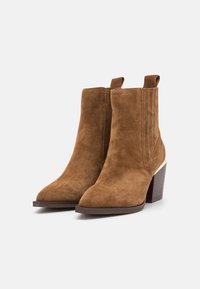 Un paio di stivaletti alla caviglia in suede marrone con tacco a blocco impilato e pannelli elastici laterali su uno sfondo bianco.