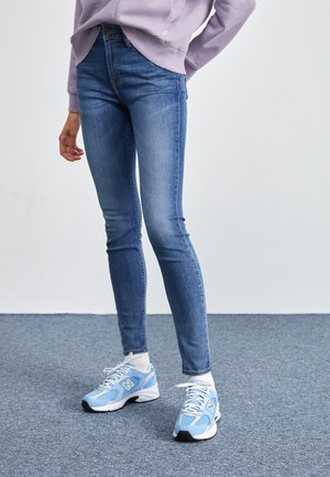 Person wearing blue skinny jeans, white socks, and light blue New Balance sneakers standing on a gray carpeted floor.