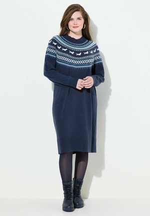 Marineblauwe gebreide jurk met lange mouwen, voorzien van een geborduurd bovenstuk in blauw en wit. Geribbelde manchetten en zoom, gecombineerd met zwarte veterschoenen.