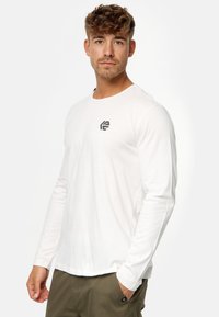 Weißes Langarmshirt mit rundem Halsausschnitt und einem kleinen schwarzen Logo auf der Brust. Der Stoff wirkt glatt und hat eine lockere Passform.