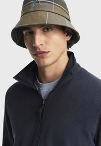 Giacca a zip in pile blu navale con colletto alto; sovrapposta a una maglietta azzurra chiara; completata con un cappello a secchiello a quadri marroni con un logo discreto.