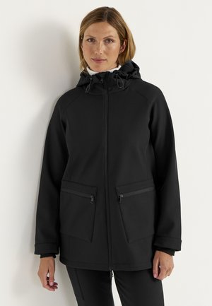Kvinde med kort hår iført en sort hættejakke med lynlås og to store lynlåslommer foran, står op ad en hvid væg.