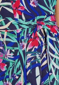 Květinové šaty z látky s námořnickým pozadím, živými květy v červené a fialové barvě, a látkovým opaskem uvázaným v pase s hladkým povrchem.