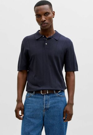 Mann iført en mørk navy kortermet poloshirt stukket inn i blå denimjeans med et brunt belte mot en ensfarget bakgrunn.