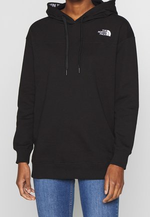 Osoba nosząca czarną bluzę The North Face z kapturem i sznurkami oraz niebieskie dżinsy, pokazana od połowy klatki piersiowej do bioder na jednolitym tle.
