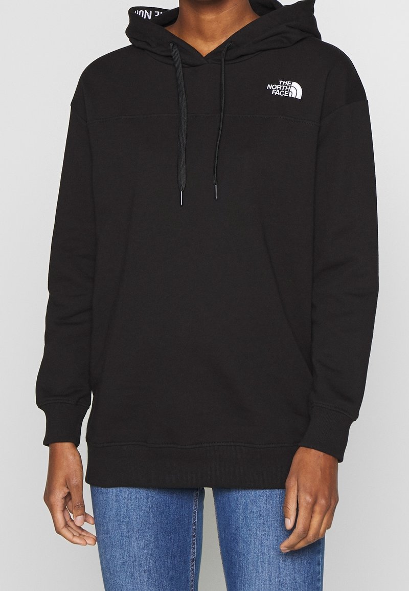 Personne portant un sweat à capuche noir The North Face avec cordons et un jean bleu, visible de la poitrine aux hanches, sur un fond uni.