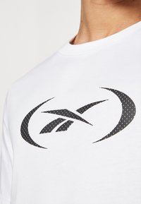 Hvit bomulls T-skjorte med et stort svart logo med kurvede linjer og prikkede mønstre. Klassisk rund hals design, korte ermer.