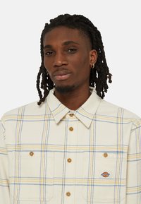 Camisa de franela para hombre en color crema con un patrón a cuadros amarillo y azul, que cuenta con dos bolsillos en el pecho y cierre de botones. Fabricada en tejido suave.