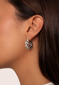 Pendientes de plata con un diseño en espiral y bordes dentados. La superficie es brillante y reflectante, destacando los detalles intrincados de la forma.