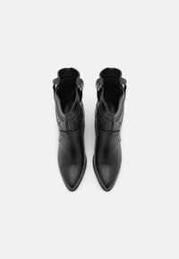 Para czarnych skórzanych botków z wąskim noskiem, sięgających do kostki, z dekoracyjnym przeszyciem i pętlami do wkładania, widocznych z góry na białym tle.