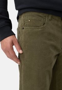 Olivgrüne Cordhosen mit strukturiertem Finish, ausgestattet mit einer Vordertasche und einem Knopf-Detail. Eine Hand ruht an der Seite.