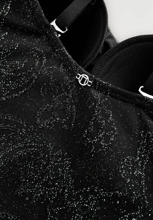 Schwarzer Stoff mit einem schimmernden, strukturierten Muster, das mit einem silbernen Akzent versehen ist und verstellbaren schwarzen Trägern ausgestattet ist. Die Innenseite ist glatt.