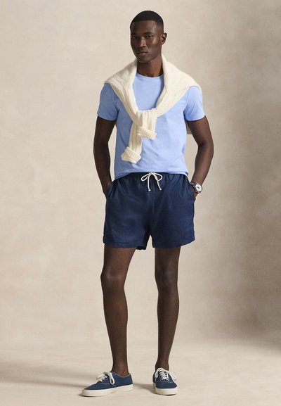 Άνδρας που φορά γαλάζιο μπλουζάκι, σκούρα μπλε σορτς με κορδόνι, κρεμ πουλόβερ περασμένο στους ώμους, σκούρα μπλε αθλητικά παπούτσια και ρολόι στον καρπό, στέκεται μέσα σε κλειστό χώρο.