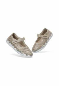 Zapatos Mary Jane perforados de color beige con suela de goma blanca y una correa en el empeine, ubicados sobre un fondo blanco liso.