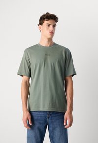 Grøn bomulds T-shirt med rund hals, korte ærmer og et præget logo på brystet. Bæret med blå denimjeans.