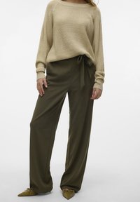 Pull en tricot beige avec un large décolleté, associé à un pantalon taille haute vert olive avec une ceinture nouée. Les chaussures sont à talons pointus avec des nœuds.