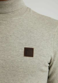 Pull à col roulé beige en maille avec un petit patch carré marron arborant un motif noir minimaliste sur la poitrine.