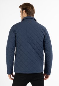 Ein Mann trägt eine marineblaue Steppjacke, steht mit dem Rücken zur Kamera, hat schwarze Hosen und kurze braune Haare vor einem weißen Hintergrund.