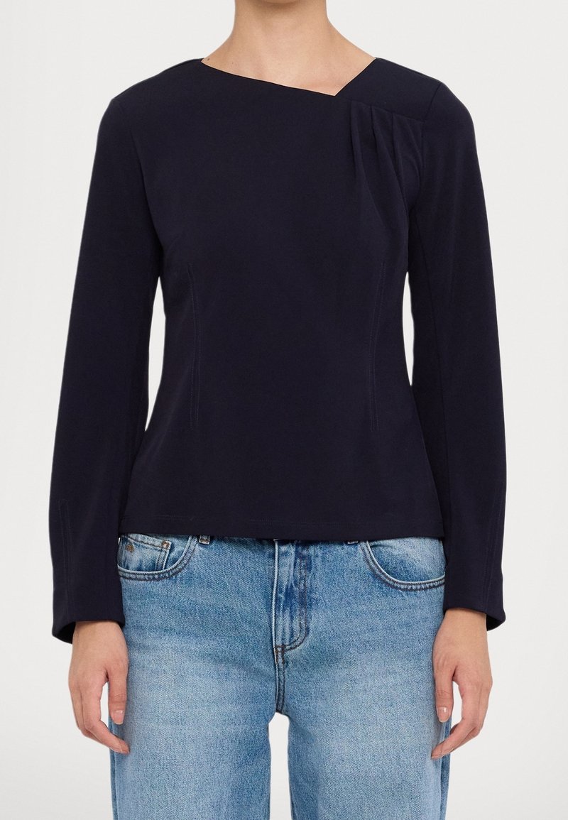 Haut à manches longues bleu marine avec encolure asymétrique, détail plissé subtil sur l'épaule et texture lisse. Associé à un jean bleu clair.