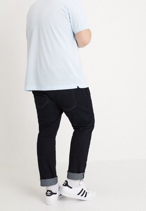 Personne portant un t-shirt bleu clair, un jean foncé retroussé, des baskets blanches Adidas avec des rayures noires, debout devant un fond blanc uni.