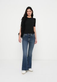 Μαύρο κοντομάνικο τοπ με υφή και φουσκωτούς ώμους, συνδυασμένο με μπλε καμπανέ τζιν και λευκά αθλητικά παπούτσια, σε στυλ με αξεσουάρ από φτερά.