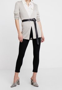 Cardigan lavorato a maglia grigio chiaro con maniche lunghe, fasciato in vita con una cintura in pelle nera, abbinato a leggings neri e mule con tacco texturizzato.